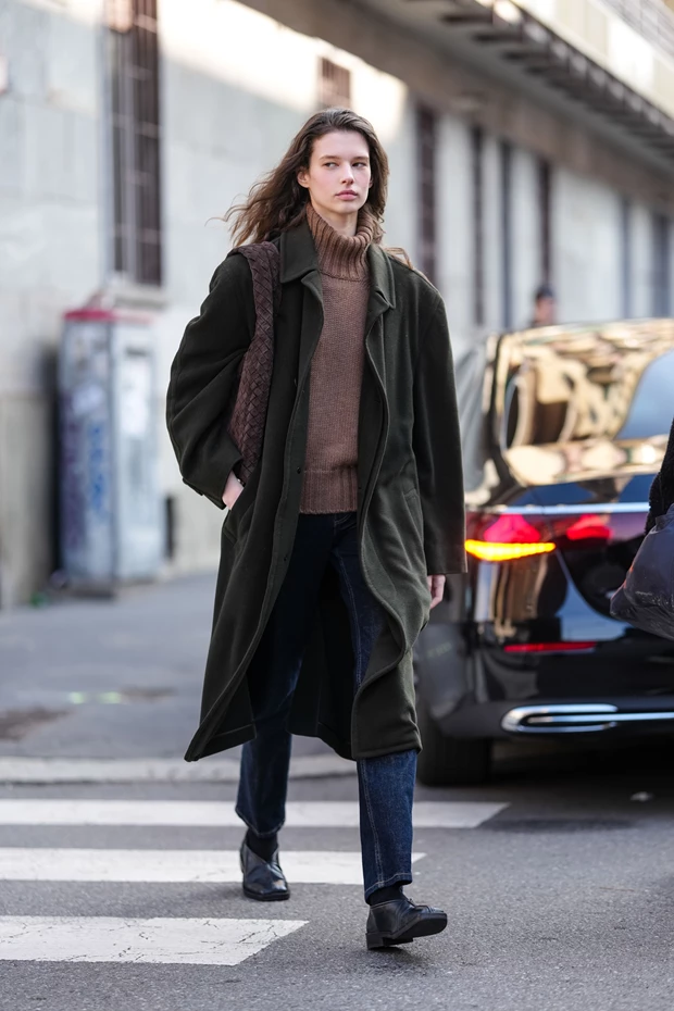 Γυναίκα με καφέ ζιβάγκο πουλόβερ, σκούρο πράσινο μακρύ παλτό, τζιν και μαύρα μποτάκια.