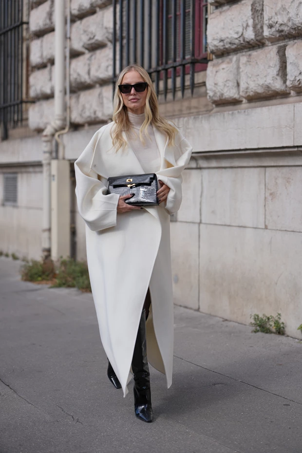 Γυναίκα με μακρύ λευκό παλτό και μαύρη τσάντα, φορώντας μαύρες ψηλές μπότες από λουστρίνι.