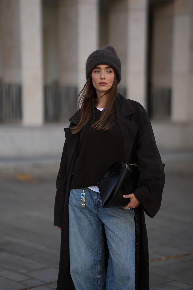 Γυναίκα με σκούρο καφέ πουλόβερ V-neck, ανοιχτόχρωμο φαρδύ τζιν, μακρύ σκούρο παλτό και γούνινο σκούρο σκούφο.