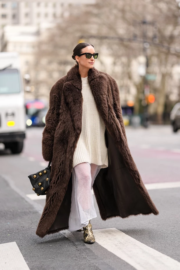 Γυναίκα στο street style που φοράει φαρδύ πουλόβερ με λευκή μάξι φούστα, γούνινο παλτό και statement ankle boots σε χρυσό.