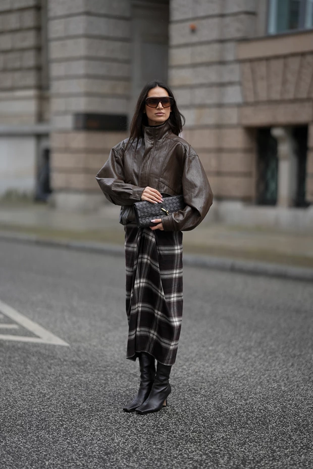 Γυναίκα με καφέ δερμάτινο μπουφάν, καρό μίντι φούστα σε καφέ-λευκές αποχρώσεις και σκούρες μπότες με τακούνι.