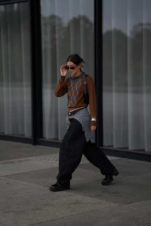 Γυναίκα με cropped καφέ πουλόβερ argyle, πολύ φαρδύ μαύρο τζιν και ογκώδη μποτάκια, περπατά στο δρόμο.