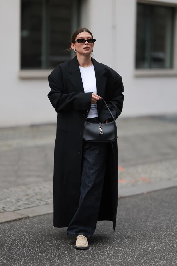 Γυναίκα με μακρύ μαύρο παλτό και σκούρο τζιν, κρατώντας μαύρη τσάντα και φορώντας μπεζ loafers.