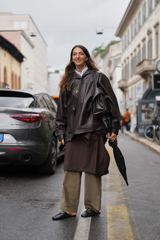 Minimal μαύρα δερμάτινα slip-on clogs σε βρεγμένο δρόμο, συνδυασμένα με λαδί παντελόνι και καφέ δερμάτινο τζάκετ