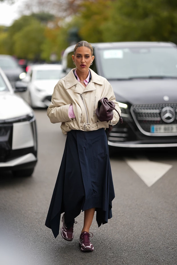 Γυναίκα που φοράει μπεζ cropped τζάκετ, ασύμμετρη μίντι φούστα σε μπλε και sneakers σε μοβ χρώμα. Κρατάει clutch.