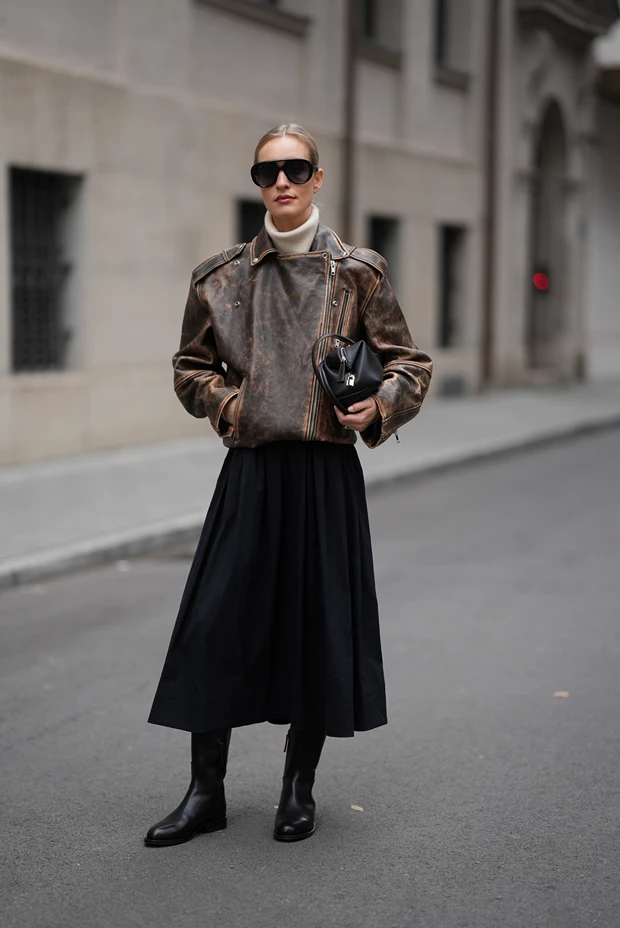 Γυναίκα με καφέ vintage δερμάτινο τζάκετ, λευκό ζιβάγκο, μαύρη μίντι φούστα και μαύρες δερμάτινες μπότες.