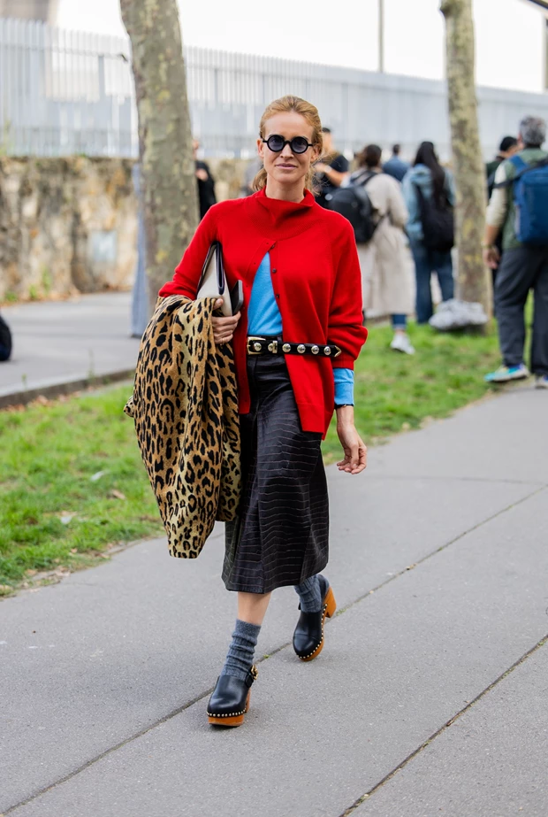 Γυναίκα με κόκκινη πλεκτή ζακέτα, γαλάζιο μπλουζάκι, μαύρη φούστα μέχρι το γόνατο, μαύρη ζώνη με διακοσμητικές λεπτομέρειες και ένα λεοπάρ παλτό διπλωμένο στο χέρι της. Φοράει γκρι κάλτσες και μαύρα clogs.