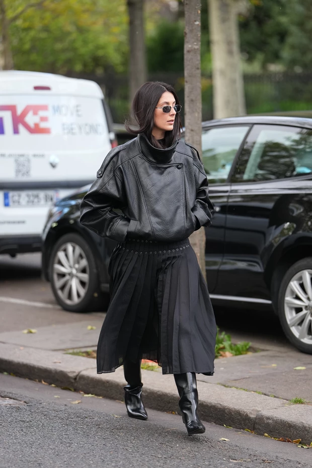 Γυναίκα με μαύρο δερμάτινο τζάκετ, μαύρη πλισέ μίντι φούστα και μαύρες ψηλές μπότες σε στυλ street style.