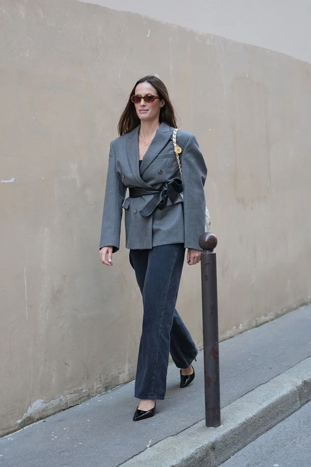 Γυναίκα με γκρι σακάκι δεμένο με ζώνη και σκούρο τζιν, φορώντας μαύρες μυτερές γόβες από λουστρίνι.