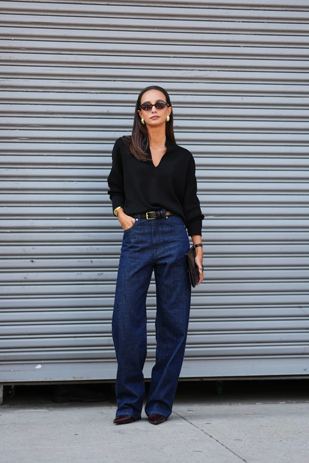 Γυναίκα με μίνιμαλ στιλ. Φοράει μαύρη μπλούζα, τζιν παντελόνι, μυτερές γόβες, λεπτή μαύρη ζώνη, χρυσά σκουλαρίκια και γυαλιά ηλίου.