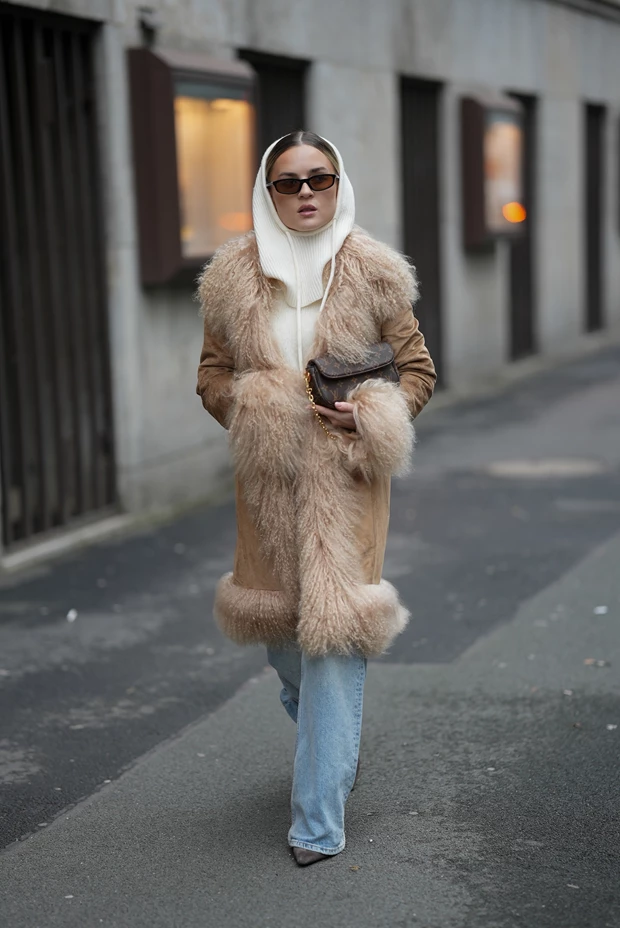 Γυναίκα φοράει ένα σουέτ παλτό σε ταμπά απόχρωση με πλούσια γούνινη επένδυση στον γιακά και στα μανίκια, συνδυασμένο με λευκό πλεκτό balaclava.