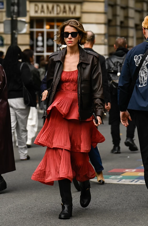 Street style εμφάνιση με κόκκινο μίντι φόρεμα με πλισέ βολάν, καφέ δερμάτινο τζάκετ, μαύρες μπότες και γυαλιά ηλίου.