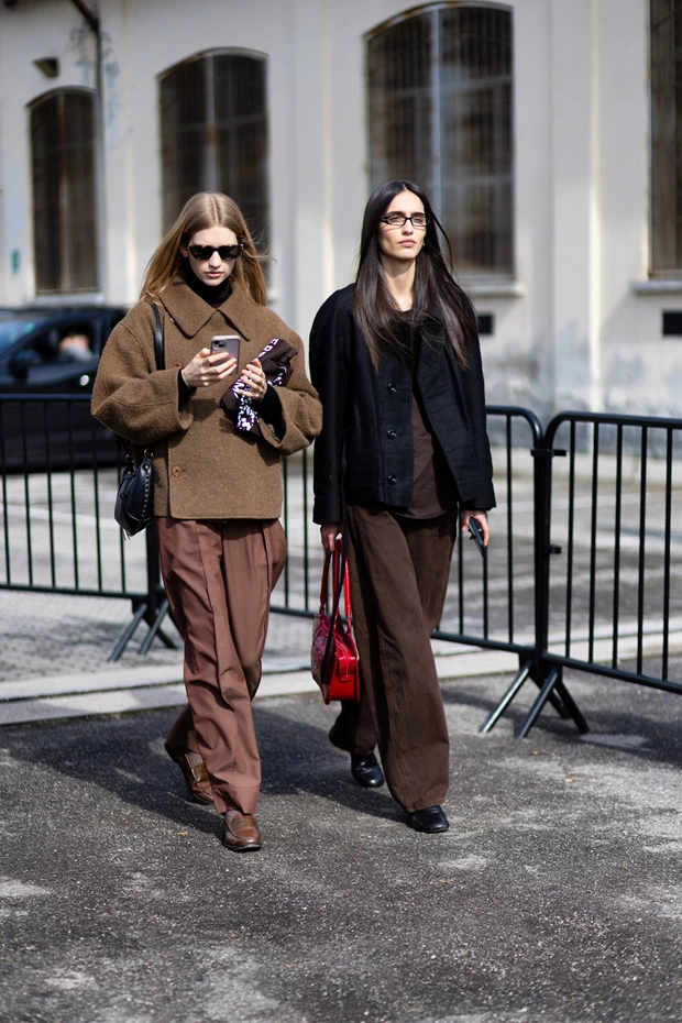 Street style: Δύο γυναίκες περπατούν φορώντας μονοχρωματικά, ουδέτερα σύνολα, που περιλαμβάνουν καφέ παντελόνια και σκούρα καφέ ή μαύρα σακάκια/τζάκετ, υποδεικνύοντας μια capsule γκαρνταρόμπα.