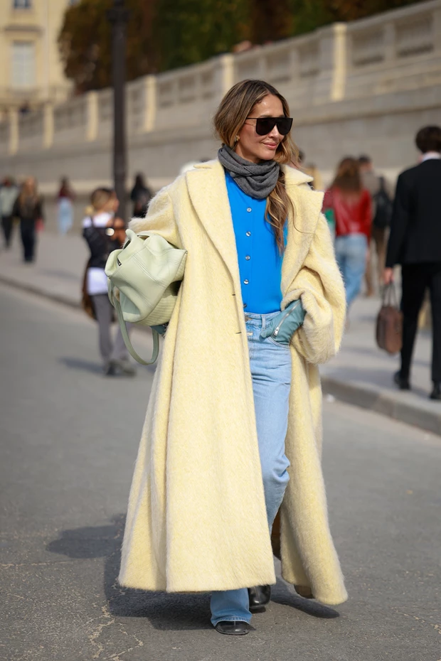 Γυναίκα με μακρύ, χνουδωτό, απαλό κίτρινο παλτό, μπλε ζακέτα με κουμπιά, τζιν παντελόνι και γκρι μάλλινο κασκόλ. Κρατάει μια πράσινη τσάντα και φοράει γυαλιά ηλίου.