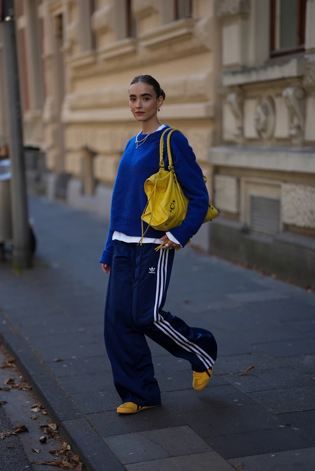 Γυναίκα με παντελόνι φόρμας, μπλε πουλόβερ και κίτρινα sneakers, σε street style look ιδανικό για το φθινόπωρο.