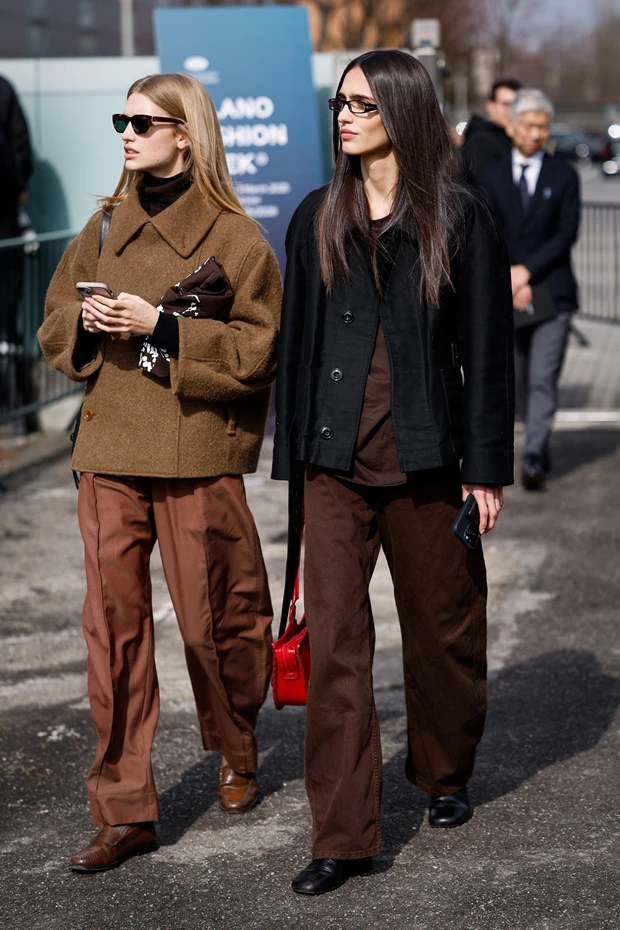 Δυο γυναίκες σε street style. H αριστερή φοράει καφέ τζάκετ, καφέ ζιβάγκο, καφέ παντελόνι και loafers, η δεξιά φοράει  μαύρο τζάκετ, καφέ σοκολατί πουκάμισο, καφέ τζιν και μαύρα loafers, ενώ κρατάει κόκκινη τσάντα.