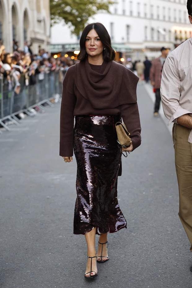 Γυναίκα σε εμφάνιση street style με μακριά φούστα με μπορντό παγιέτες. Τη συνδυάζει με καφέ πλεκτό πουλόβερ και μαύρα πέδιλα.