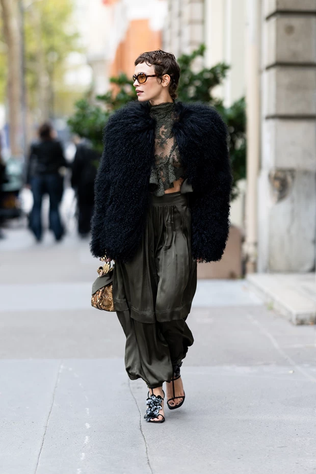 Γυναίκα φοράει λαδί φόρεμα με βολάν και δαντέλα με shearling παλτό σε μαύρο χρώμα και με πέδιλα. Κρατάει animal print τσάντα.