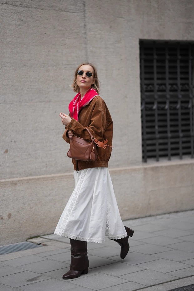 Γυναίκα με καφέ suede bomber jacket, λευκό μίντι φόρεμα και καφέ μπότες.