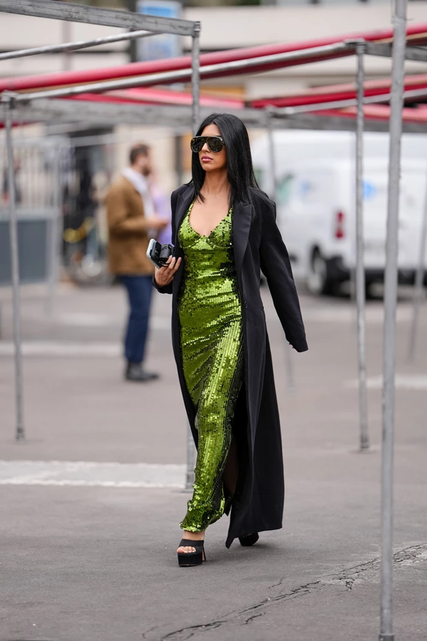 Γυναίκα με μακρύ μαύρο παλτό πάνω από λαχανί φόρεμα με παγιέτες και μαύρα πέδιλα.
