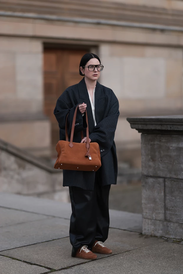 Γυναικείο street style με oversized σκούρο παλτό, μαύρο παντελόνι, καφέ sneakers και καφέ tote τσάντα σε χειμερινό outfit.