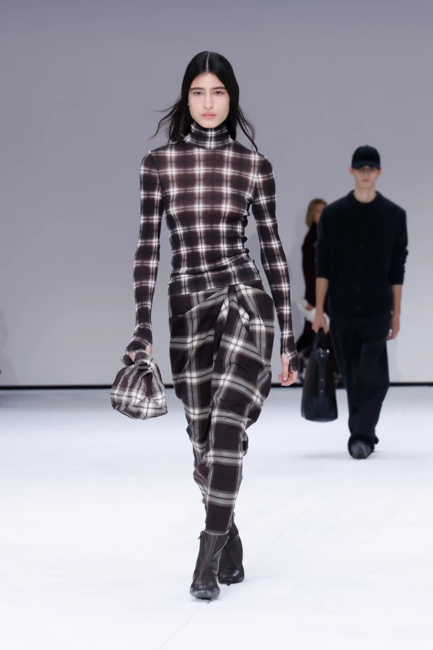 Στιγμιότυπο από την πασαρέλα της Cos για τη συλλογή Φθινόπωρο - Χειμώνας 2025: καρό φούστα με καρό ζιβάγκο, μποτάκια και καρό clutch.