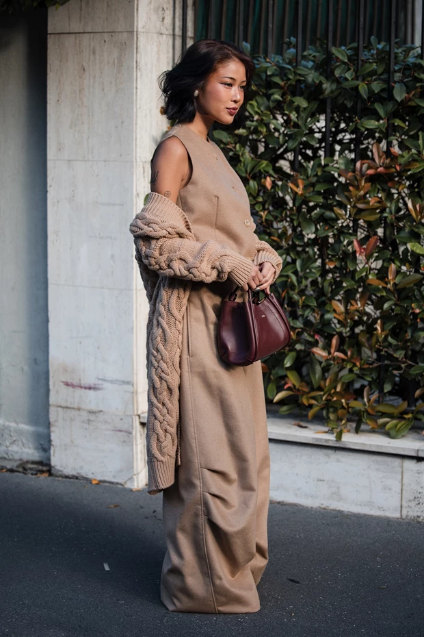 Κομψή γυναίκα με μακριά μπεζ πλεκτή ζακέτα ριγμένη στους ώμους της, φορεμένη πάνω από ένα μπεζ γιλέκο και παντελόνι. Κρατάει μια μπορντό τσάντα.