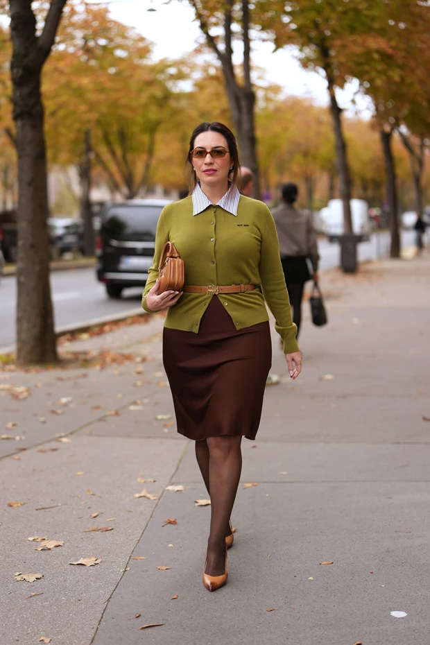 Γυναίκα με πράσινη ζακέτα και καφέ φούστα περπατά, φορώντας ταμπά γυαλιστερές γόβες.