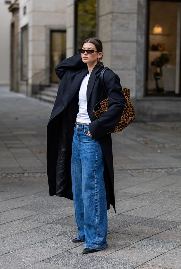 Γυναίκα που φοράζει τζιν, λευκό T-shirt και μαύρο παλτό. Κρατά λεοπάρ τσάντα.