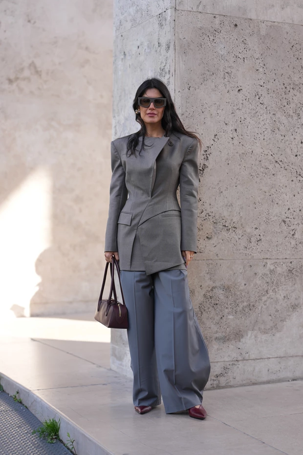Γυναίκα με γκρι σακάκι και φαρδύ παντελόνι, κρατώντας τσάντα και φορώντας μπορντό παπούτσια με μυτερή μύτη.