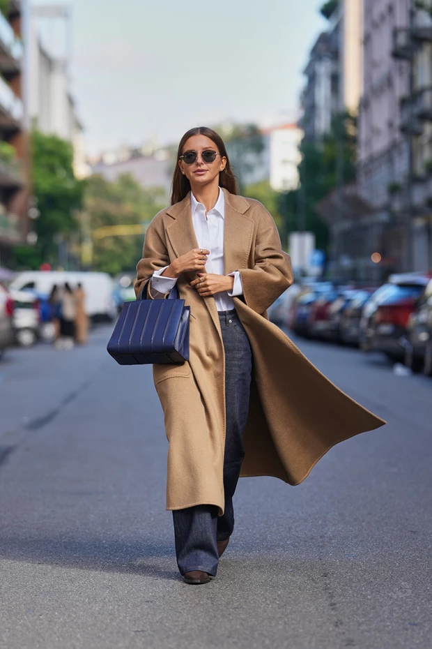 Γυναίκα στον δρόμο που φοράει τζιν παντελόνι, πουκάμισο και καμηλό παλτό, ενώ κρατά τσάντα σε μπλε χρώμα.