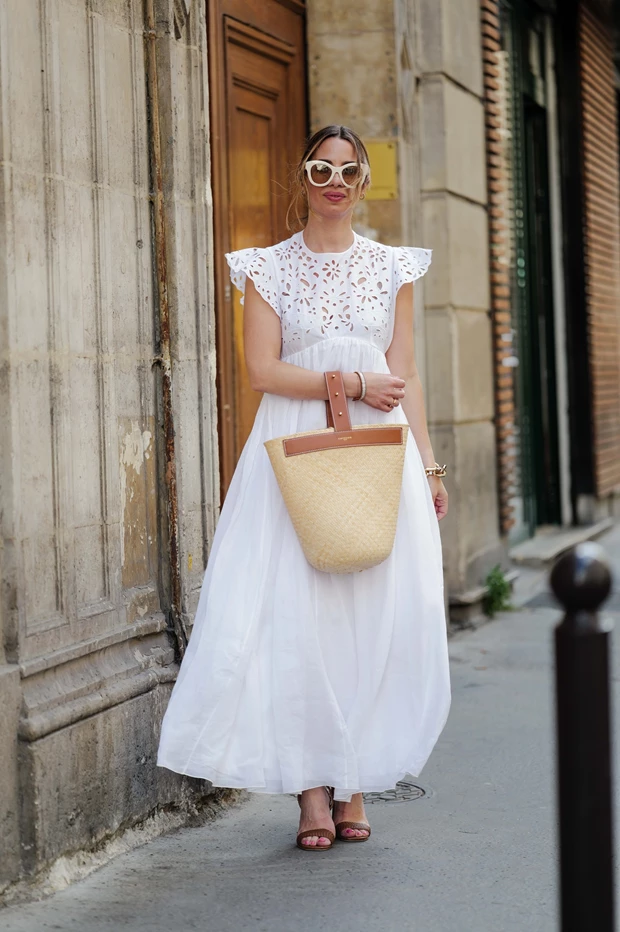 Γυναίκα με λευκό midi φόρεμα με broderie λεπτομέρειες και raffia τσάντα, καλοκαιρινό street style με φυσικές υφές και ουδέτερους τόνους