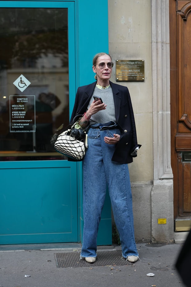 Γυναίκα με φαρδύ τζιν παντελόνι και ναυτικό μπλε σακάκι, φορώντας ανοιχτόχρωμα μπεζ/κρεμ flat παπούτσια.