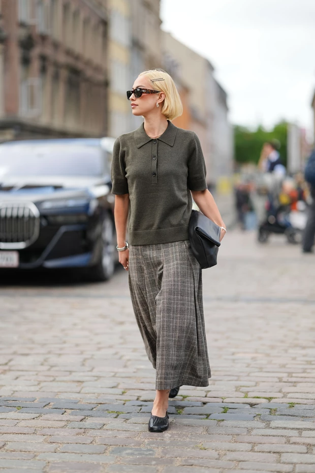 Γυναίκα με σκούρο πράσινο πλεκτό polo μπλουζάκι και μακριά πλισέ καρό φούστα, περπατά σε πλακόστρωτο δρόμο.