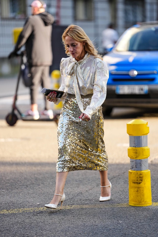 Γυναίκα με χρυσή μίντι φούστα με παγιέτες, συνδυασμένη με λευκή μεταξωτή μπλούζα με φουσκωτά μανίκια, φιόγκο στον λαιμό και λευκές γόβες.