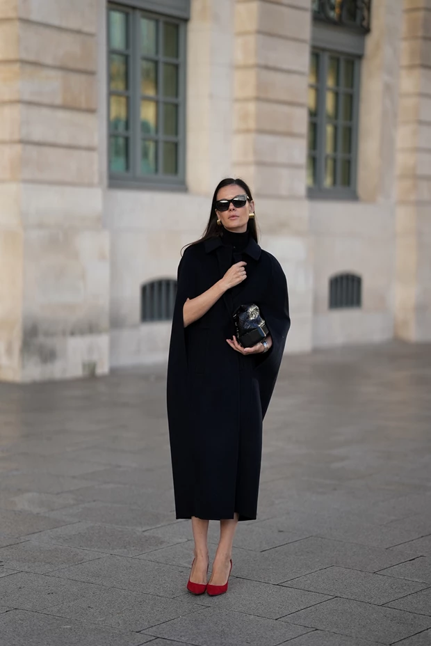 Σικάτο μαύρο midi φόρεμα-κάπα με κόκκινες γόβες και χρυσά σκουλαρίκια, επίσημο ντύσιμο γραφείου για τις γιορτές 2025.