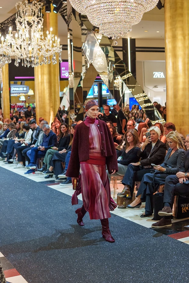 Μοντέλο με μπορντό-ροζ layered σύνολο, που αποτελείται από παλτό, ζιβάγκο και πλισέ φούστα, στην πασαρέλα του Golden Hall.