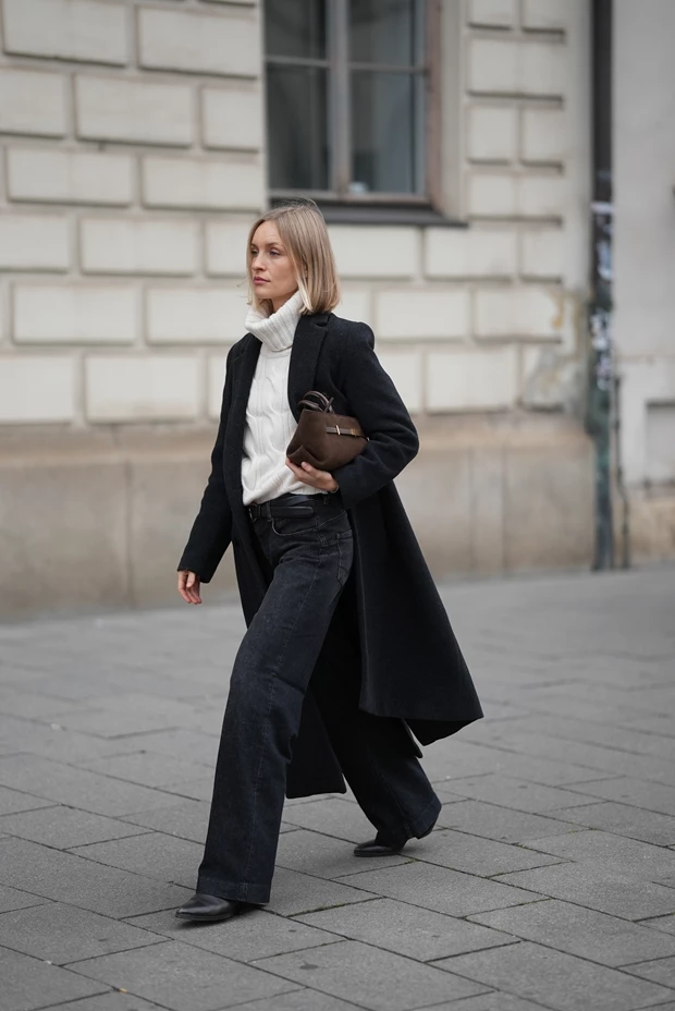 Γυναίκα με λευκό πλεκτό πουλόβερ ζιβάγκο, μαύρο φαρδύ τζιν, μακρύ μαύρο παλτό και καφέ τσάντα.