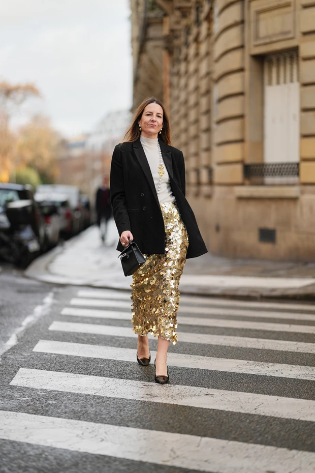 Γυναίκα περπατάει φορώντας μίντι φούστα με χρυσές παγιέτες. Συνδυάζει τη φούστα με ένα ζιβάγκο και ένα μαύρο σακάκι