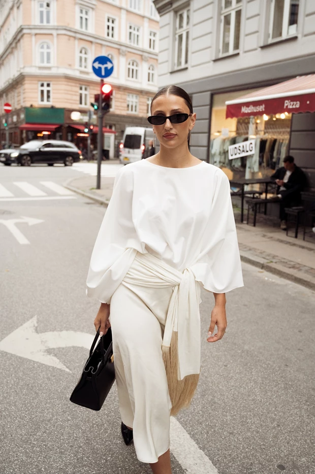 Γυναίκα με total white σύνολο με loose μπλούζα και draped midi φούστα με δέσιμο στη μέση, minimal street style quiet luxury με μαύρη structured τσάντα