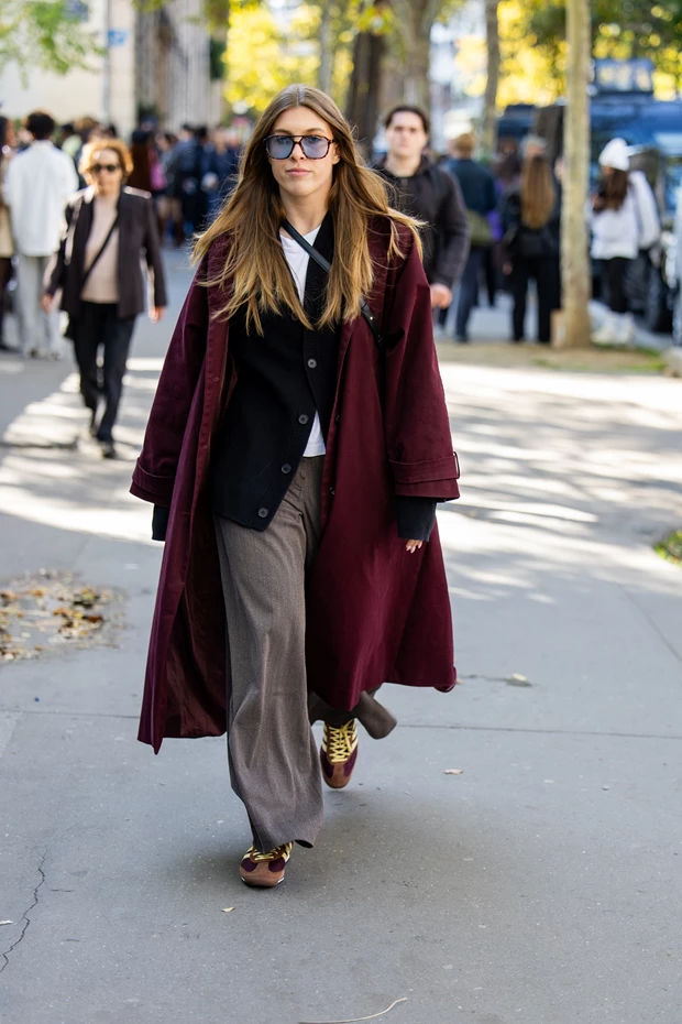 Γυναίκα με μακρύ μπορντό παλτό, μαύρη ζακέτα, λευκό t-shirt και φαρδύ γκρι-καφέ παντελόνι. Φοράει σκούρα γυαλιά ηλίου και κίτρινα-μπορντό sneakers.