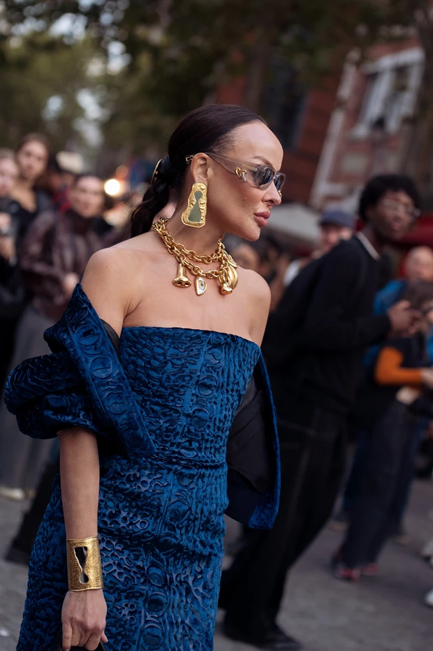 Γυναίκα με μπλε στράπλες φόρεμα, ογκώδη χρυσά κοσμήματα και γυαλιά ηλίου.