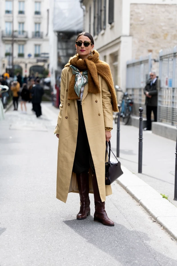 Γυναίκα με μπεζ καμπαρντίνα, καφέ κασκόλ, μεταξωτό φουλάρι και καφέ μπότες στέκεται σε πλακόστρωτο δρόμο.