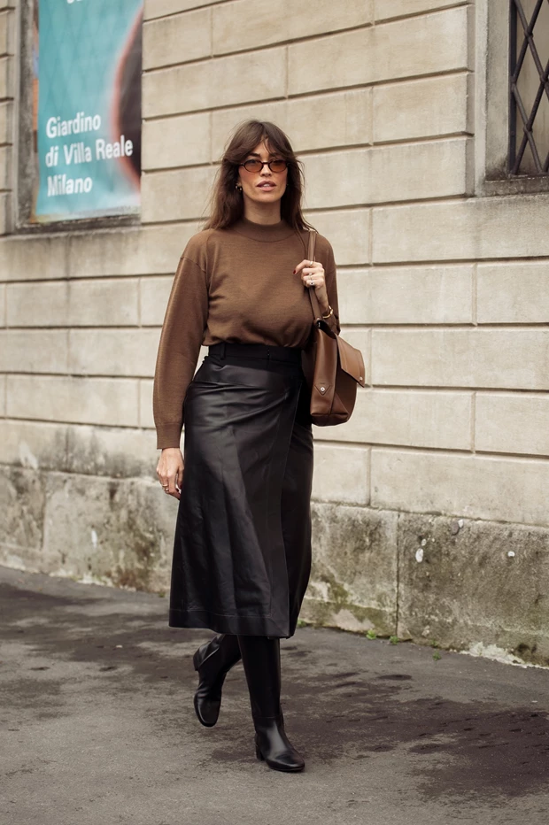 Γυναίκα με καφέ πουλόβερ, μαύρη δερμάτινη μίντι φούστα τύπου wrap και μαύρες ψηλές μπότες.