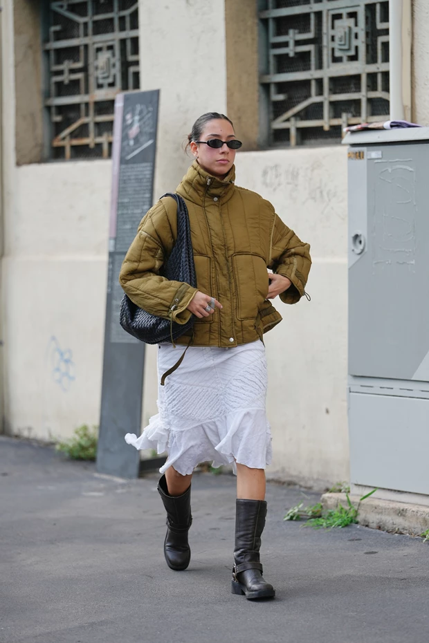 Μια γυναίκα φοράει ένα κοντό, χακί/καφέ puffer μπουφάν, λευκή φούστα, μαύρες μπότες και γυαλιά ηλίου, κρατώντας μια μεγάλη πλεκτή τσάντα.