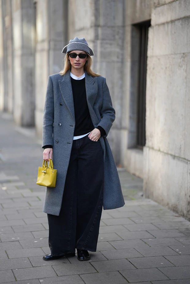 Street style εμφάνιση με πολύ φαρδύ μαύρο barrel-leg τζιν, γκρι μακρύ παλτό, κίτρινη μικρή τσάντα και πλεκτό καπέλο.