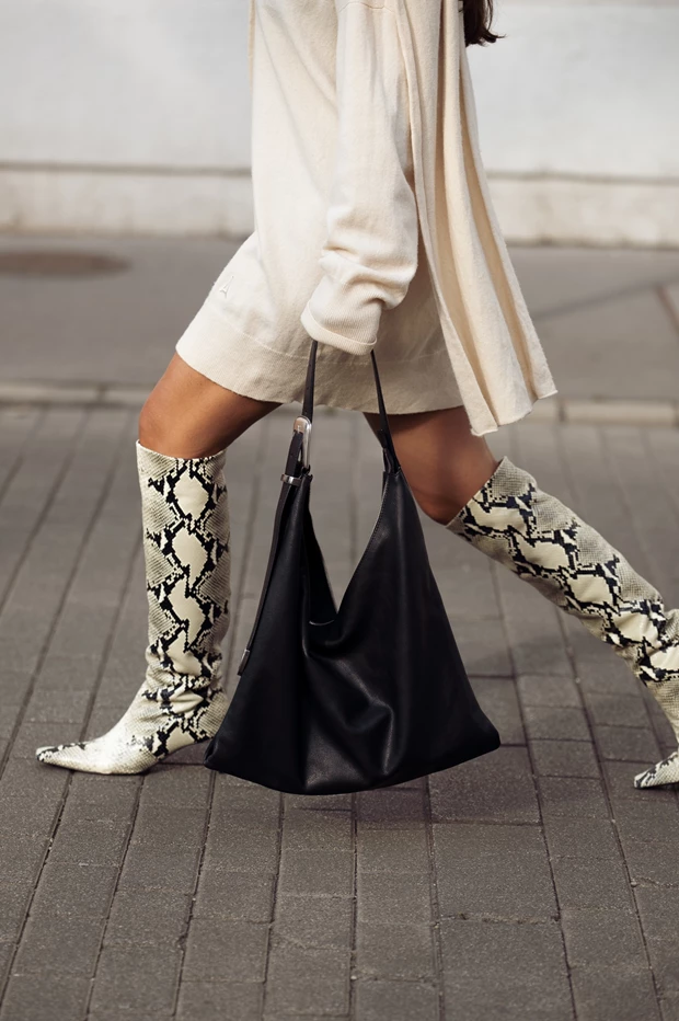 Γυναίκα περπατά φορώντας ψηλές μπότες με σχέδιο δέρματος φιδιού, μπεζ φόρεμα και κρατώντας μαύρη δερμάτινη τσάντα.