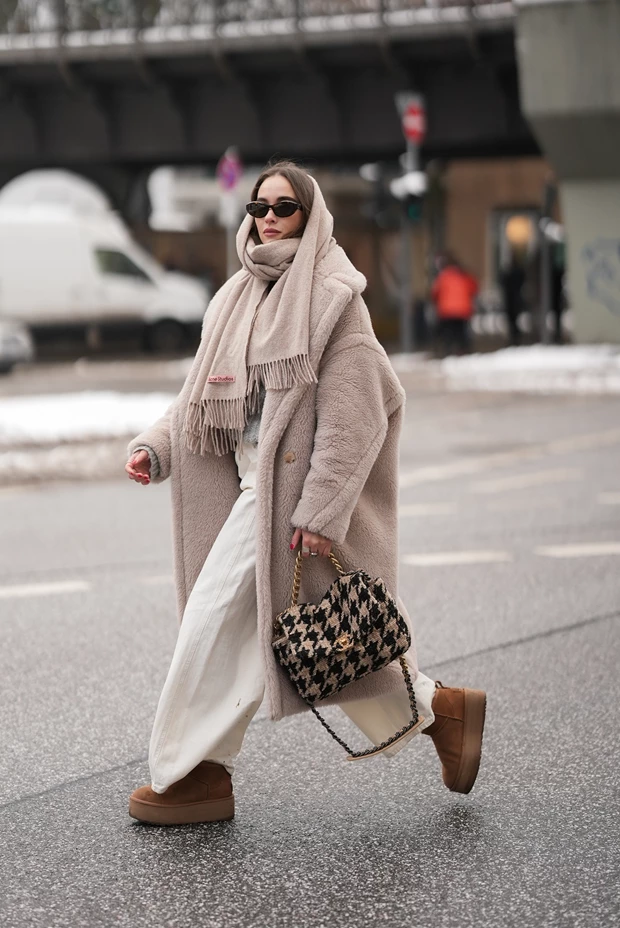 Γυναίκα στο χιόνι με λευκό παντελόνι, fluffy παλτό, κασκόλ τυλιγμένο στο κεφάλι και γούνινα μποτάκια.